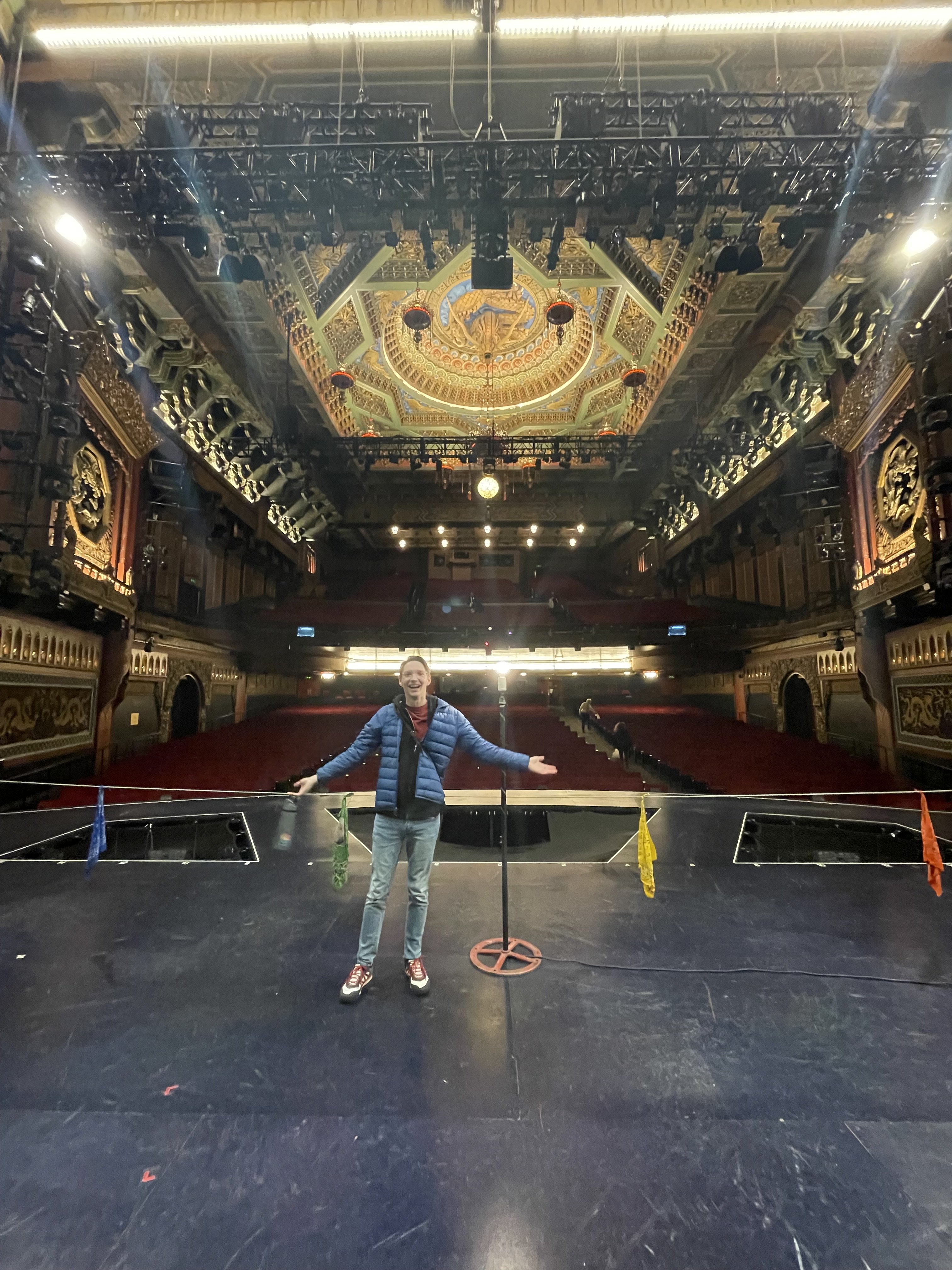 Nicholas Main on stage in a grand ornate theatre, arms open wide